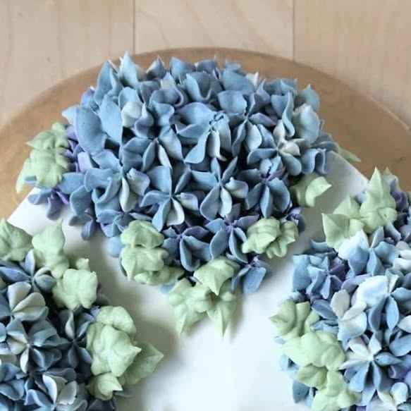 Cake topped with buttercream hydrangeas in blue, purple and white with pale green leaves